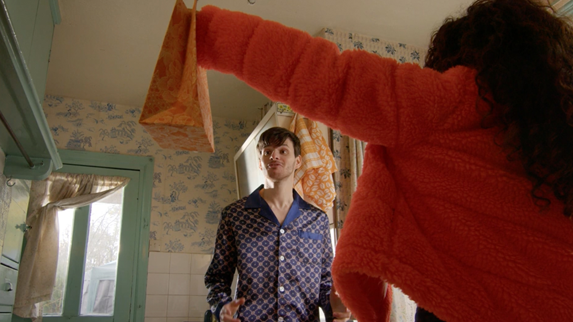 Sam Hume in blue pyjamas stares at a present being held by an outstretched arm wearing a fluffy orange jacket